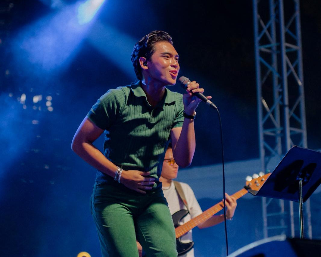 Man (rayyanroulette) singing into a microphone on stage with blue lights and equipment in the background